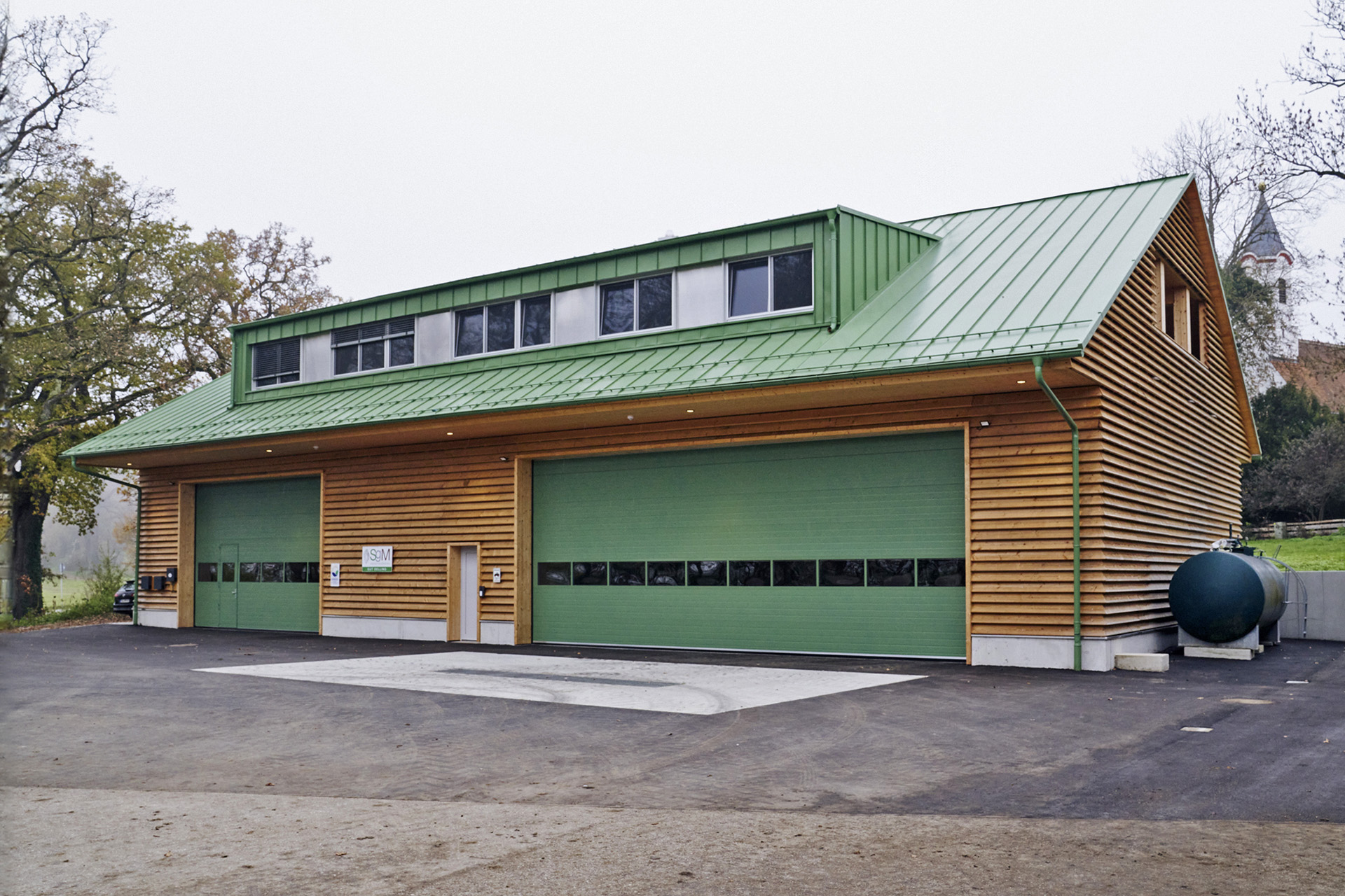 Kategorie Öffentliche Projekt: ein langgezogenes Wohn- und Gewerbegebäude mit horizontaler Holzfassade, grünem Blechdach und Gauben sowie grünen Garagentoren